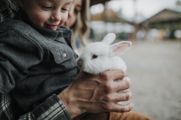 Nahaufnahme von niedlichen lächelnden Sohn spielen mit Kaninchen von Eltern auf dem Bauernhof - CAVF62827