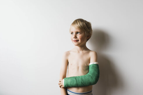 Thoughtful shirtless boy with broken arm looking away while standing against wall at home - CAVF62832