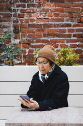 Ältere Frau in warmer Kleidung, die ihr Smartphone benutzt, während sie auf einer Bank vor einer Backsteinmauer in der Stadt sitzt - CAVF62867