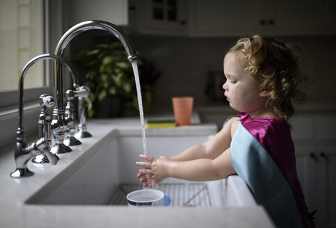 Side view of girl wearing pink cape washing hands in kitchen sink while standing at home - CAVF62871