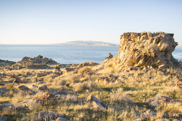 Blick aus mittlerer Entfernung auf eine Frau, die auf einer Felsformation sitzt und auf das Meer blickt - CAVF62885