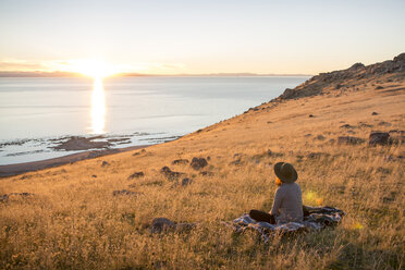 Rear view of woman looking at sea while sitting on grassy field against sky during sunset - CAVF62886