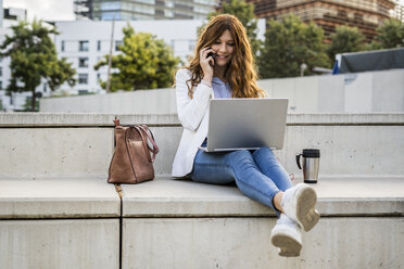 Young businesswoman sitting on bench in the city, using laptop, talking on the phone - GIOF05782