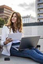 Young businesswoman sitting on stairs in the city, working with laptop, holding document - GIOF05784