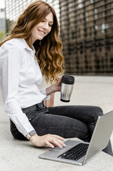 Young businesswoman sitting on bench in the city, working with laptop, drinking coffee - GIOF05824