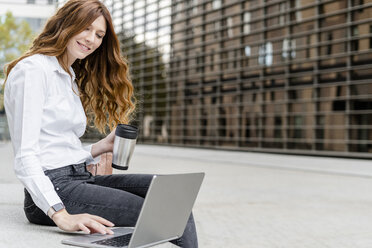 Young businesswoman sitting on bench in the city, working with laptop, drinking coffee - GIOF05825
