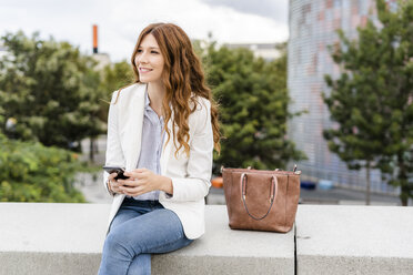 Young businesswoman commuting in the city, using smartphone, sitting on a wall - GIOF05843