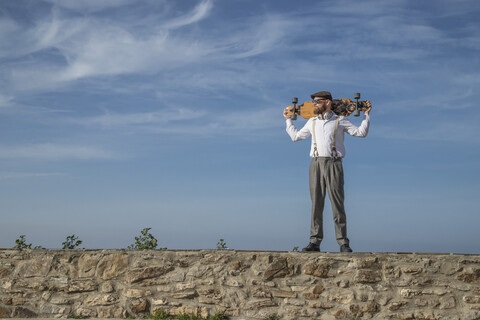 Mann mit Longboard auf den Schultern steht auf einer Mauer und schaut in die Ferne, lizenzfreies Stockfoto