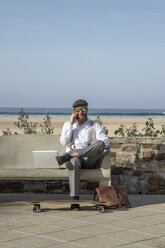 Lächelnder Mann am Telefon sitzt mit Laptop auf einer Bank vor Strand und Meer - KBF00582