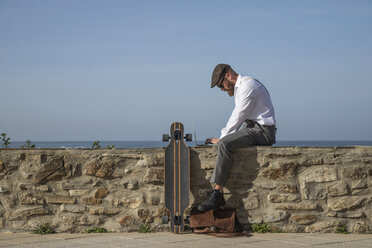 Mann sitzt auf einer Mauer vor dem Meer und arbeitet an einem Laptop - KBF00584
