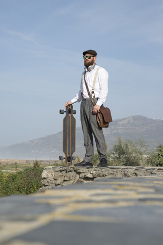 Bärtiger Mann, der mit Laptop, Longboard und Ledertasche an einer Wand steht, lizenzfreies Stockfoto