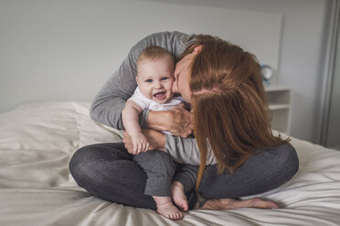Happy mother kissing cute son while sitting on bed against wall at home - CAVF62910