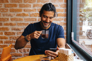 Smiling mature man with black coffee using smart phone against brick wall in cafe - CAVF62949