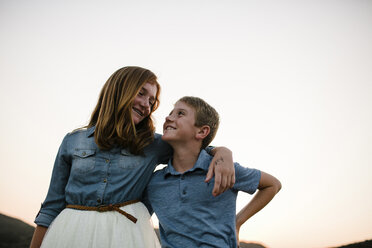 Low angle view of smiling siblings looking each other face to face while standing against clear sky during sunset - CAVF62971