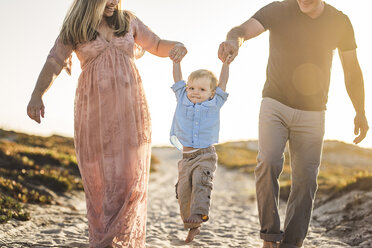 Glückliche Eltern halten die Hände ihres Sohnes, während sie am Strand gegen den klaren Himmel bei Sonnenuntergang spazieren gehen - CAVF62985