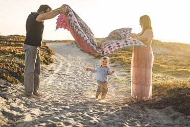 Seitenansicht der Eltern halten Decke über niedlichen Sohn läuft auf Sand am Strand gegen klaren Himmel bei Sonnenuntergang - CAVF62986