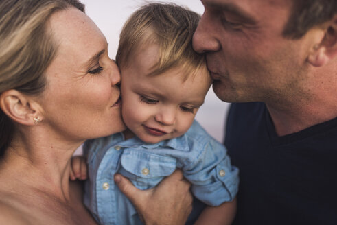 Nahaufnahme von Eltern, die ihren süßen Sohn küssen, während sie bei Sonnenuntergang am Strand stehen - CAVF62992