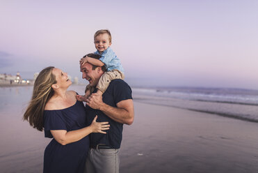 Happy husband looking at wife while carrying son on beach against sky during sunset - CAVF62993