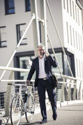 Happy mature businessman with bicycle talking on cell phone in the city - UUF16616