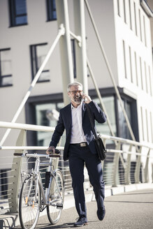 Glücklicher reifer Geschäftsmann mit Fahrrad, der in der Stadt mit einem Handy telefoniert - UUF16616