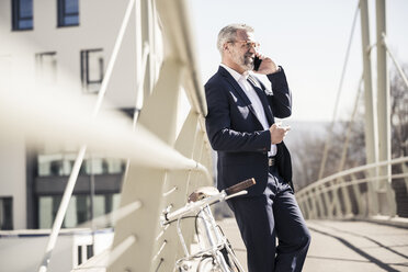 Smiling mature businessman with bicycle talking on cell phone in the city - UUF16618