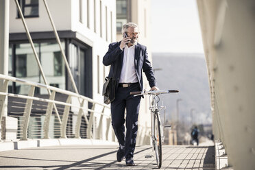 Lächelnder reifer Geschäftsmann mit Fahrrad im Gespräch am Handy in der Stadt - UUF16620