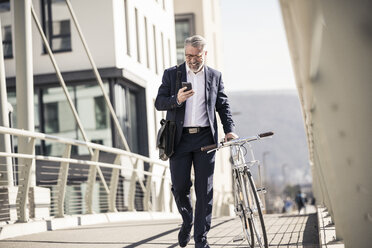 Smiling mature businessman with bicycle and cell phone in the city - UUF16621