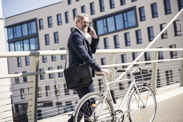Lächelnder reifer Geschäftsmann mit Fahrrad im Gespräch am Handy in der Stadt - UUF16622