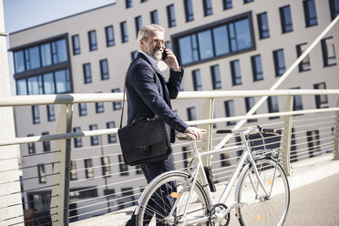 Smiling mature businessman with bicycle talking on cell phone in the city - UUF16622