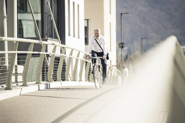 Smiling mature businessman riding bicycle on a bridge in the city - UUF16633