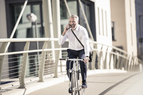 Glücklicher reifer Geschäftsmann, der mit seinem Handy telefoniert und auf einer Brücke in der Stadt Fahrrad fährt, lizenzfreies Stockfoto