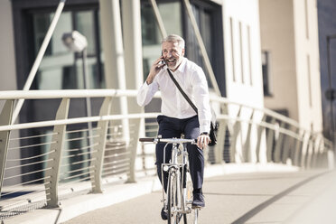 Glücklicher reifer Geschäftsmann, der mit seinem Handy telefoniert und auf einer Brücke in der Stadt Fahrrad fährt - UUF16634