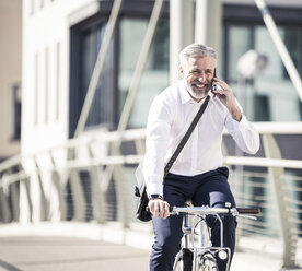 Glücklicher reifer Geschäftsmann, der mit seinem Handy telefoniert und auf einer Brücke in der Stadt Fahrrad fährt - UUF16636