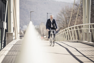 Lächelnder reifer Geschäftsmann mit Kopfhörern beim Fahrradfahren auf einer Brücke in der Stadt - UUF16637