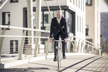 Smiling mature businessman with headphones riding bicycle on a bridge in the city - UUF16640