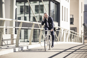 Smiling mature businessman with headphones riding bicycle on a bridge in the city - UUF16641