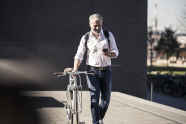 Lächelnder reifer Geschäftsmann mit Fahrrad, Mobiltelefon und Kopfhörern unterwegs - UUF16645