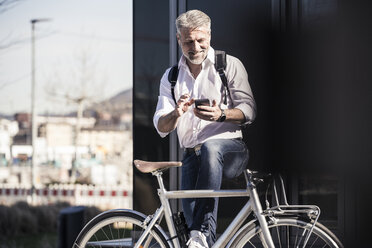 Lächelnder reifer Geschäftsmann mit Fahrrad und Kopfhörern, der ein Mobiltelefon benutzt - UUF16646