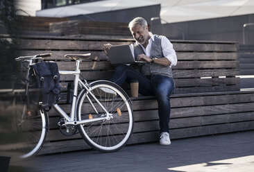 Lächelnder reifer Geschäftsmann mit Fahrrad, der auf einer Bank sitzt und einen Laptop benutzt - UUF16647