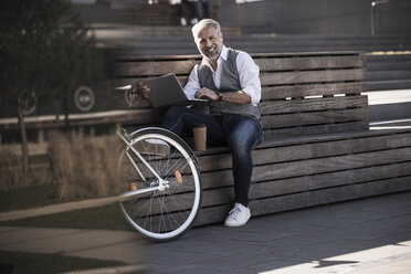 Smiling mature businessman with bicycle sitting on a bench using laptop - UUF16648
