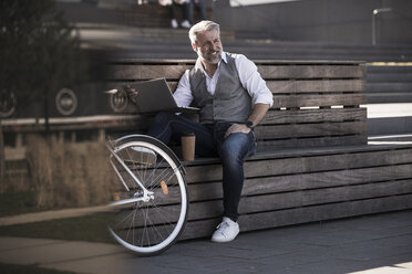 Lächelnder reifer Geschäftsmann mit Fahrrad, der auf einer Bank sitzt und einen Laptop benutzt - UUF16649