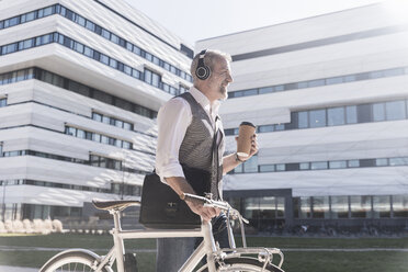 Smiling mature businessman with bicycle, takeaway coffee and headphones on the go in the city - UUF16652