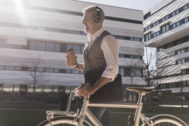 Smiling mature businessman with bicycle, takeaway coffee and headphones on the go in the city - UUF16653