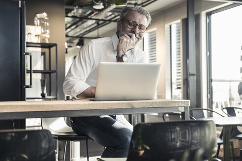 Lächelnder reifer Geschäftsmann mit Blick auf den Laptop im Büro - UUF16677