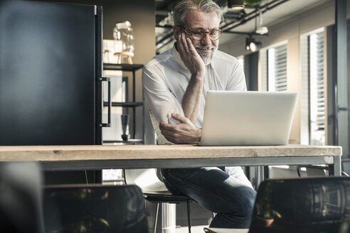 Lächelnder reifer Geschäftsmann mit Blick auf den Laptop im Büro - UUF16678