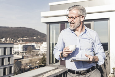 Smiling mature businessman with cup of coffee and tablet on roof terrace - UUF16689