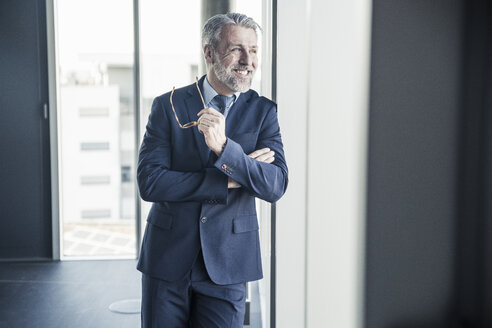 Lächelnder reifer Manager schaut aus dem Fenster im Büro - UUF16695