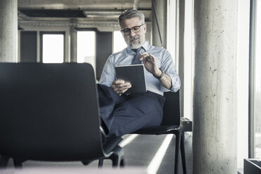 Mature businessman sitting on chair using tablet - UUF16697