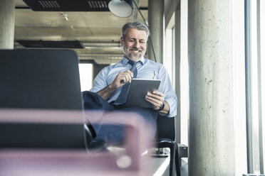 Smiling mature businessman sitting on chair using tablet - UUF16699