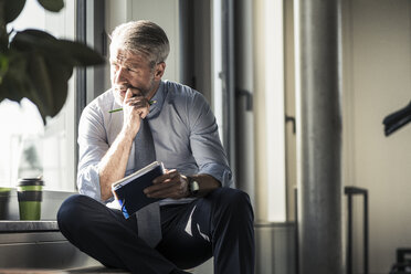 Mature businessman sitting at the window with notebook - UUF16703